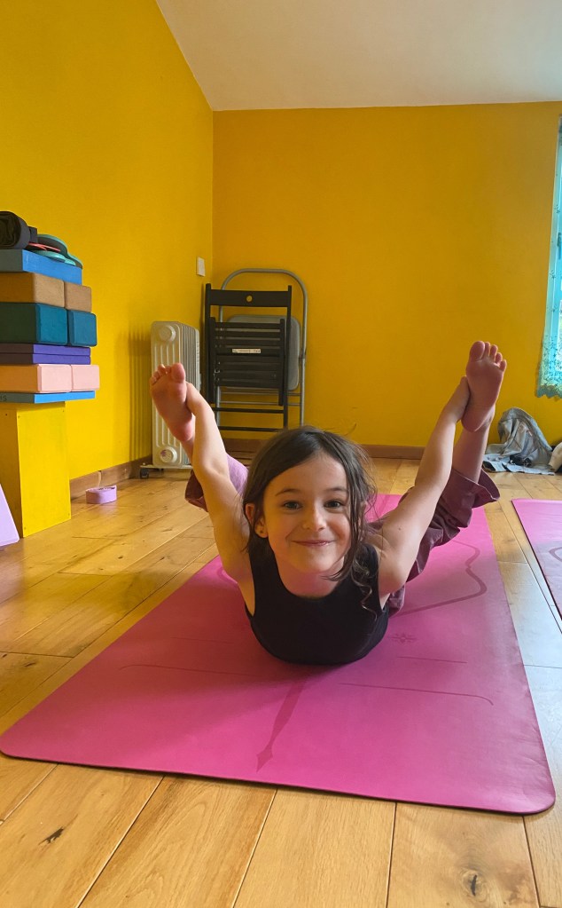 A young girl smiling while practicing a bow pose on a pink yoga mat in a bright, welcoming yoga studio with yellow walls and yoga props stacked in the background