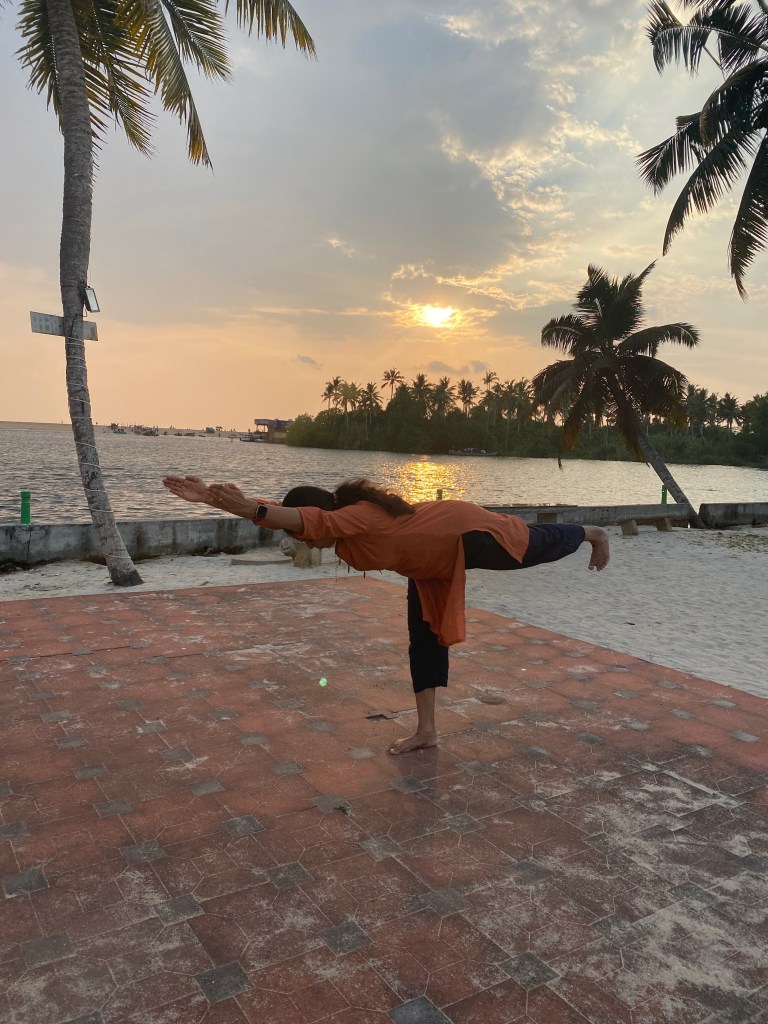 Smitha holding Warrior III pose at sunset in Trivandrum, Kerala, barefoot on red sand tiles, with palm trees and still water in the background.