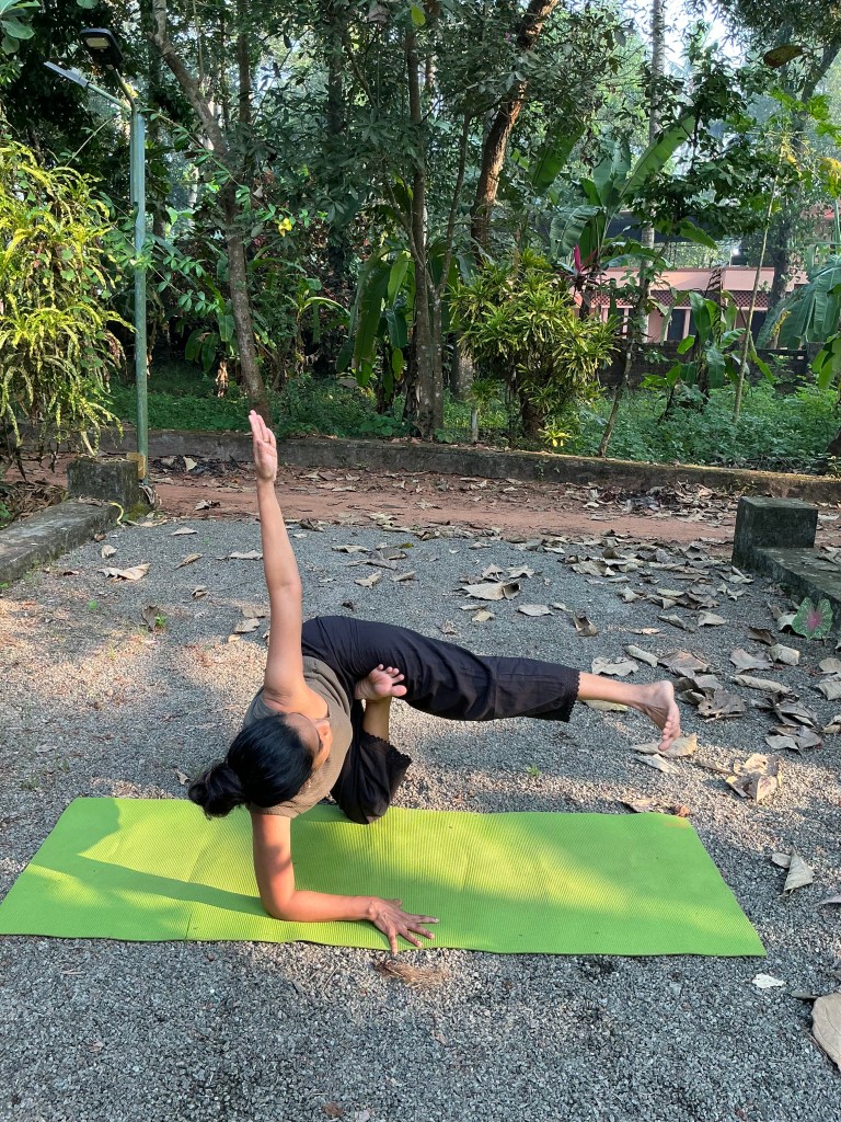 Woman practising a yoga balance pose on a green mat outdoors, surrounded by trees and greenery at a home in Kerala