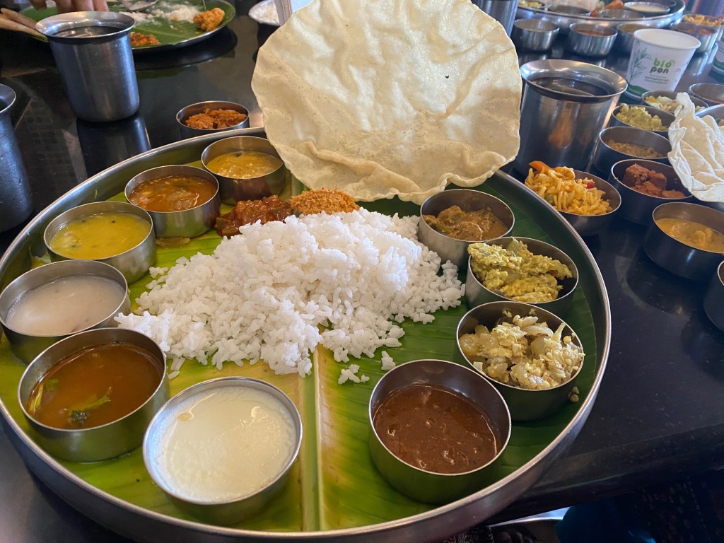 Traditional South Indian vegetarian thali served on a banana leaf with white rice, sambar, rasam, dal, vegetable dishes, pickles, papad, and payasam in small metal bowls.